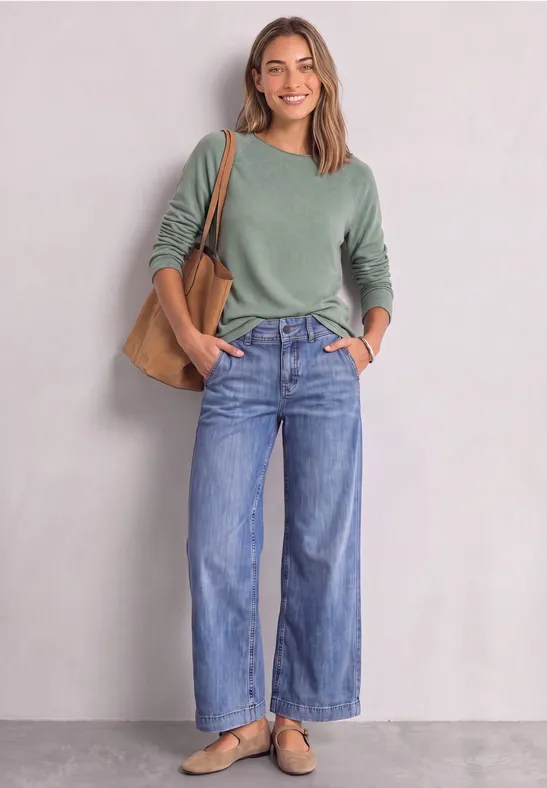 Jeune femme souriante devant un mur clair, portant un pull vert, un jean large, un sac marron et des mocassins.