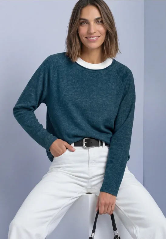 Frau sitzt vor hellem Hintergrund, trägt blaugrünen Pullover, weiße Hose und lächelt.