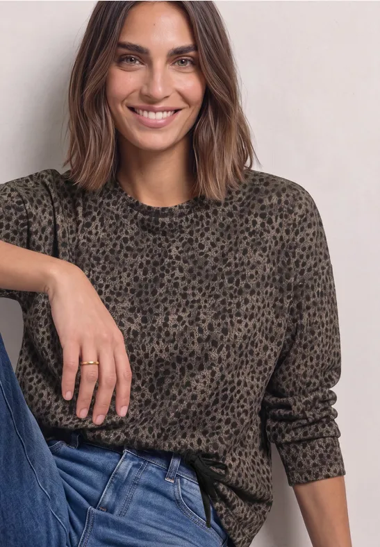 Femme aux cheveux bruns mi-longs souriante, portant un pull léopard gris-brun et un jean bleu.