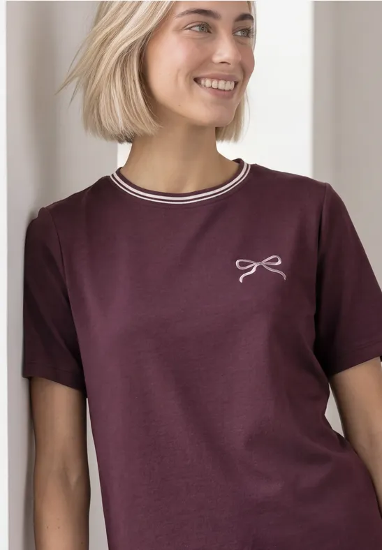 Eine junge Frau in einem burgunderroten T-Shirt mit weißem Kragenrand und kleinem Schleifen-Motiv.
