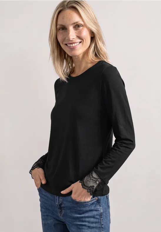 Eine lächelnde Frau mit hellem Haar trägt einen schwarzen Langarmshirt mit Spitzenbündchen und Jeans.