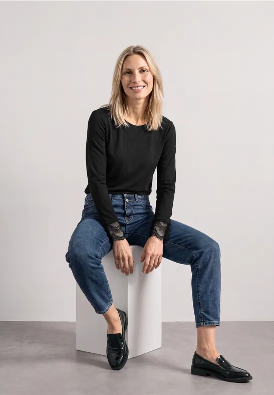 Eine blonde Frau sitzt auf einem weißen Würfel, trägt eine schwarze Bluse, Jeans und lächelt.