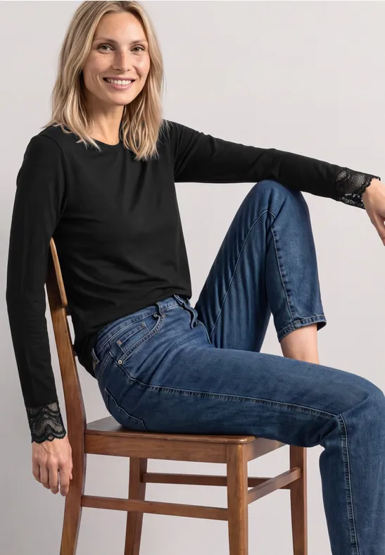 Eine lächelnde Frau in schwarzem Langarmshirt und blauen Jeans sitzt auf einem Holzstuhl vor hellem Hintergrund.