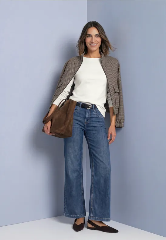 Frau mit weißem Top, weitem Jeans, taupefarbener Jacke und brauner Tasche lächelnd vor grauem Hintergrund.