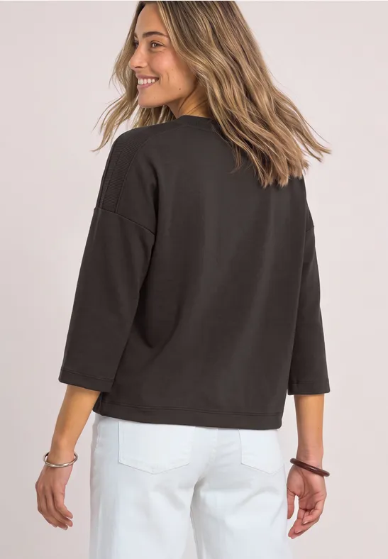 Vrouw in zwarte trui en witte broek kijkt lachend over haar schouder.