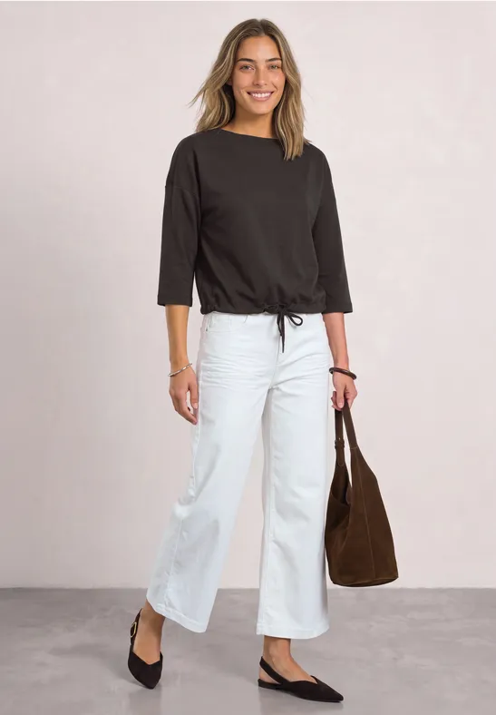 Vrouw in zwart 3/4-mouwshirt, witte wijde jeans en zwarte ballerina's, met een bruine tas.