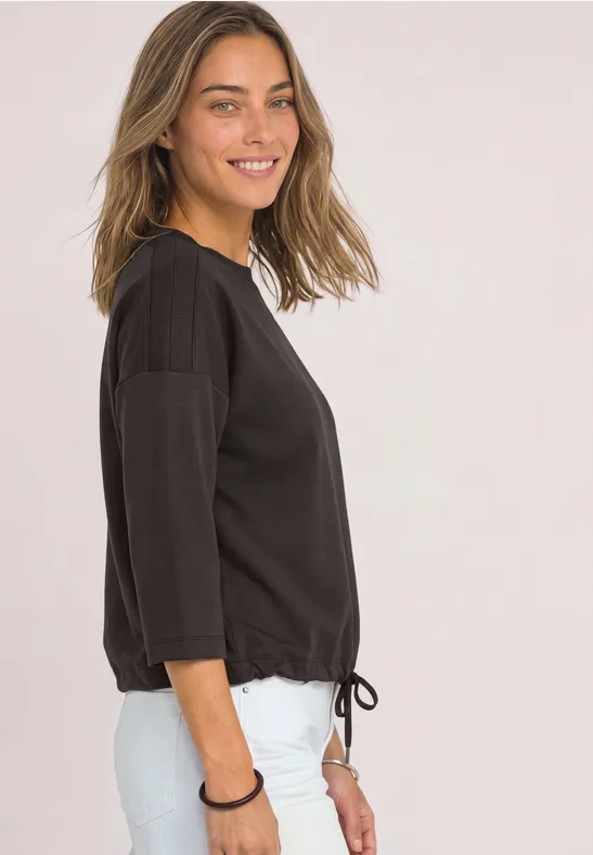 Een lachende vrouw in profiel draagt een zwarte 3/4-mouw top met strikkoord en witte broek tegen een lichte achtergrond.