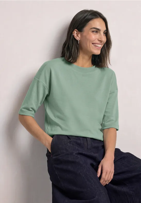 Vrouw glimlacht in een lichtgroen shirt met halflange mouwen en donkere broek tegen lichte achtergrond.