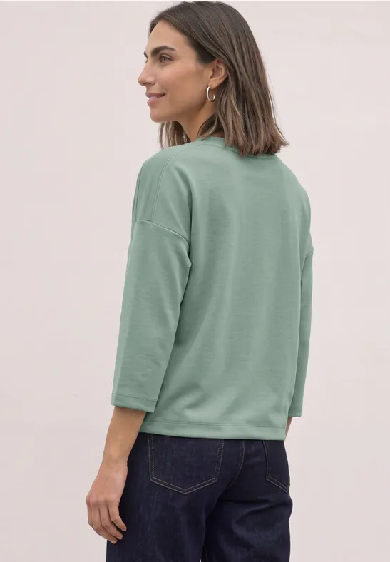 Vrouw in profiel met losse groene 3/4-mouw top en donkere spijkerbroek voor een neutrale achtergrond.