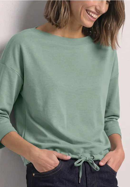 Vrouw glimlachend in een groen driekwartmouwshirt met koordje in de taille, handen in de zakken.