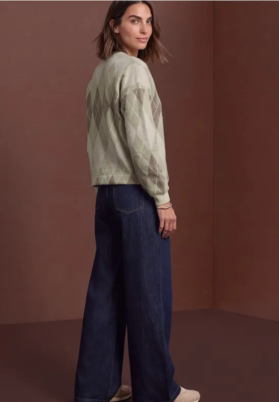 Femme en pull vert à motif, jean large et baskets blanches, debout devant un mur brun, regard par-dessus l'épaule.