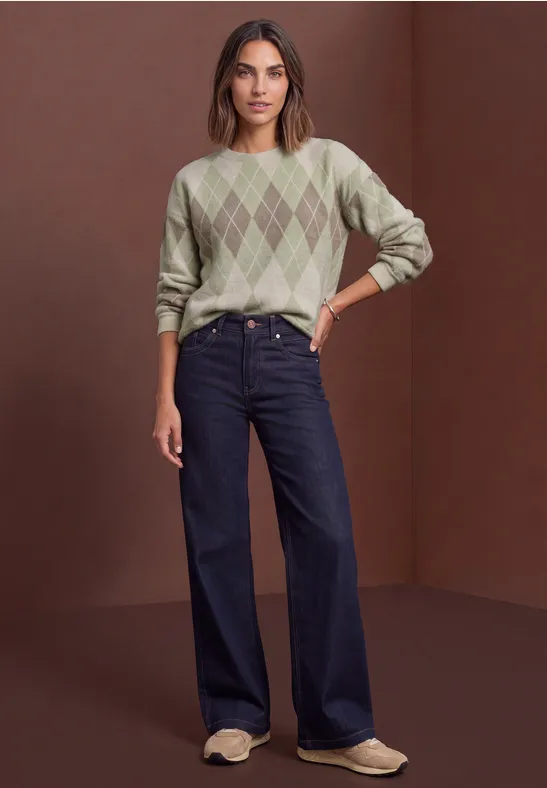 Femme en pull vert à losanges, jean large et mocassins clairs sur fond marron.