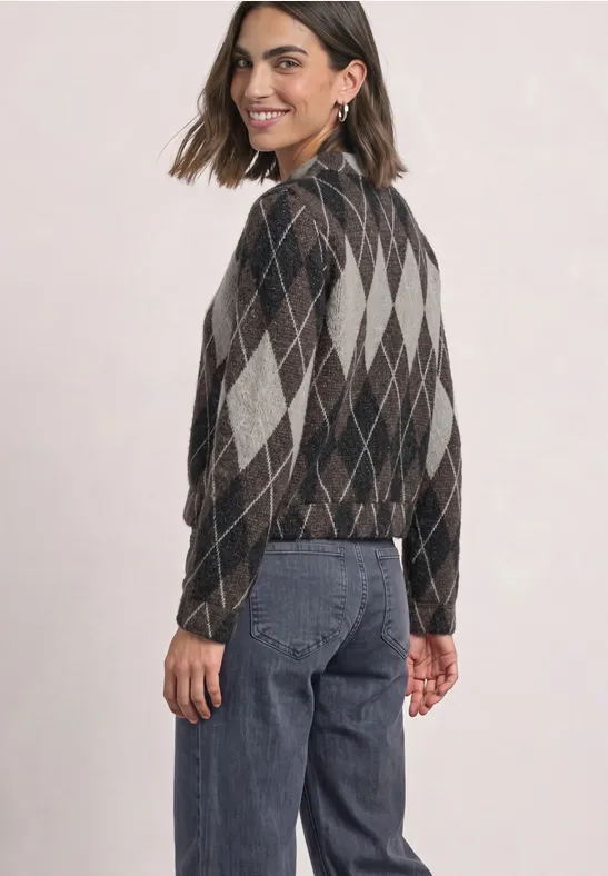 Jeune femme de profil porte un gilet à losanges et un jean, elle sourit légèrement.