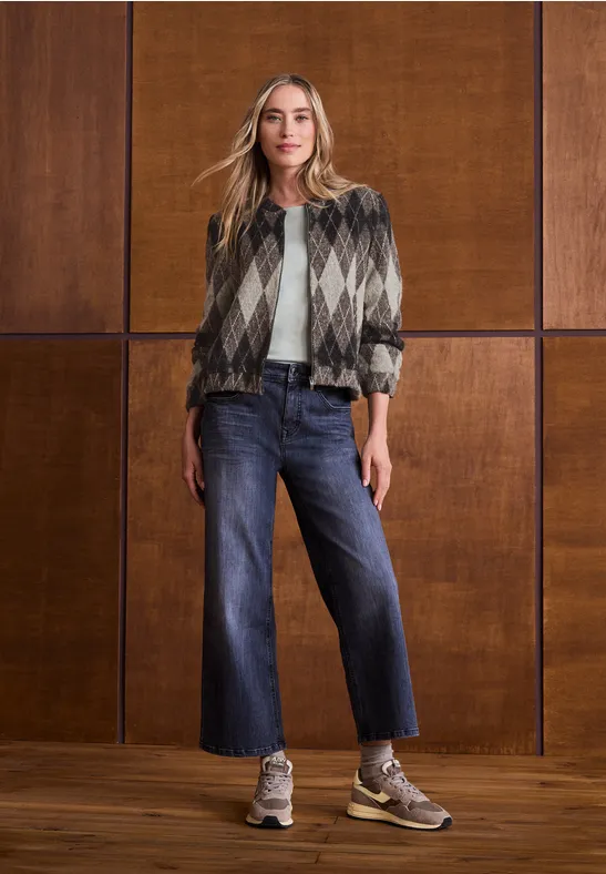 Femme portant une veste à carreaux, un t-shirt blanc, jean large et baskets devant un mur en bois.