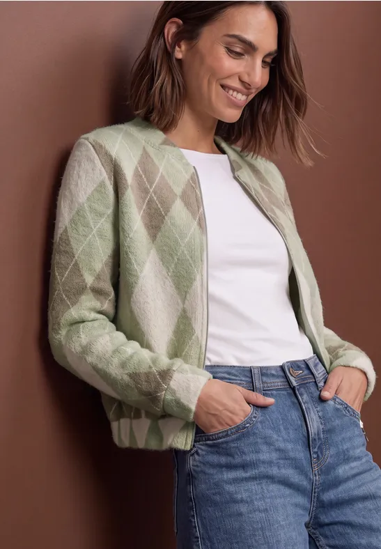 Femme souriante en cardigan losangé vert et beige, t-shirt blanc et jean bleu, mains dans les poches, fond marron.