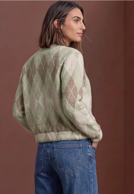 Femme vue de dos, pull à losanges vert et crème, jean bleu, devant un mur marron, regard de profil.