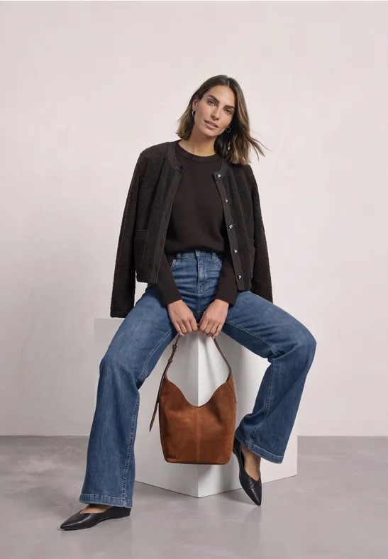 Frau sitzt auf weißem Sockel, trägt schwarzen Cardigan, Jeans und schwarze Schuhe, braune Tasche neben ihr.