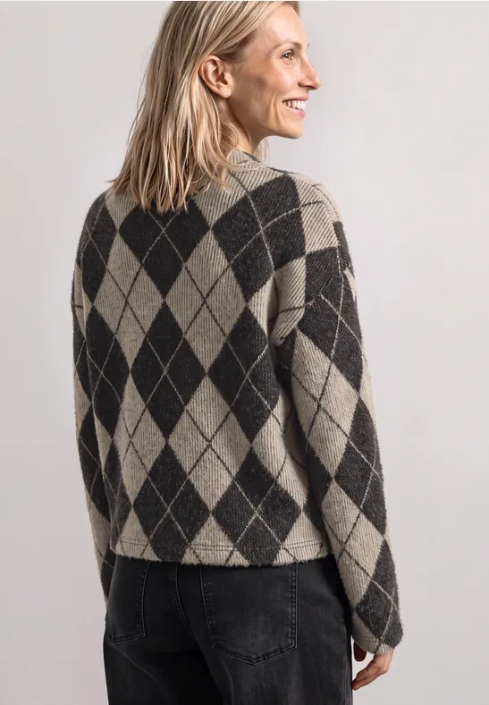 Eine lächelnde Frau mit blonden Haaren trägt einen beigen-schwarzen Rautenmuster-Pullover und Jeans.