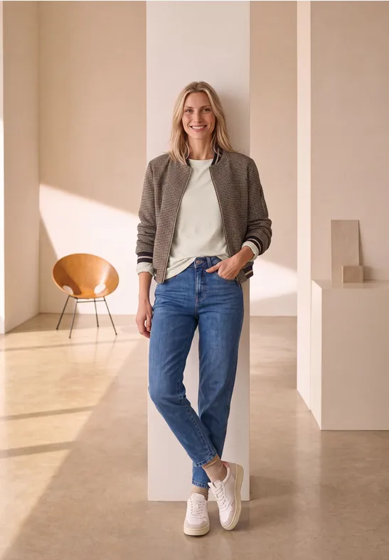 Eine lächelnde Frau in Jeans und Sneakern lehnt in einem hellen, modernen Raum an einer Säule.