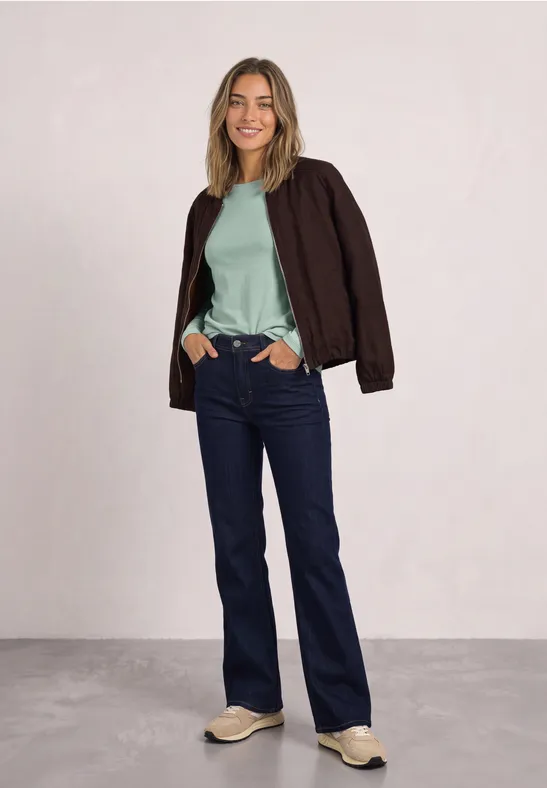 Frau in Jeans, mintgrünem Shirt, brauner Jacke und Sneakern steht lächelnd mit Händen in den Taschen vor neutralem Hintergrund.