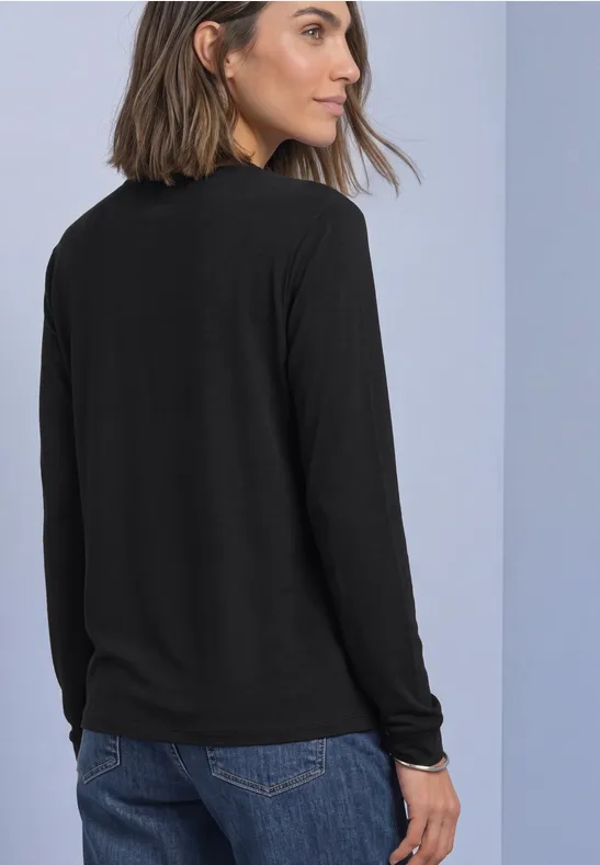 Frau mit schwarzem Langarmshirt und blauer Jeans, Rücken zur Kamera vor hellblauer Wand.
