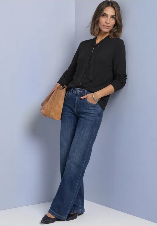 Frau lehnt an Wand; trägt schwarze Bluse, weite Jeans und halbrunde braune Tasche.