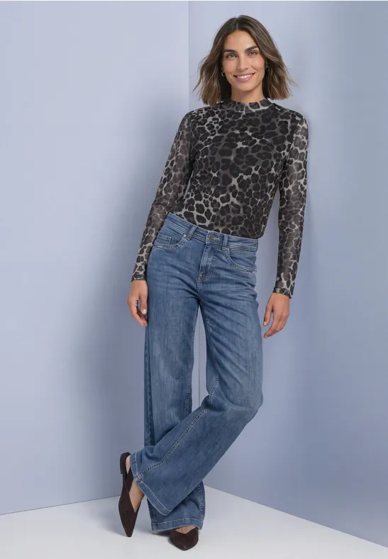 Femme appuyée contre un mur, jean large, haut à manches longues à motif et escarpins.