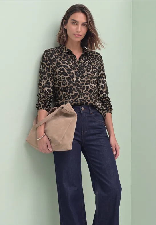 Frau in Leopardenbluse und weiten Jeans mit großer Beigetonne Handtasche vor grünem Hintergrund.
