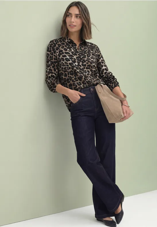 Frau lehnt lässig an einer hellgrünen Wand, trägt Leopardenbluse, weite Jeans und beige Tasche.