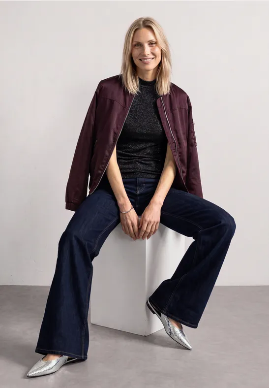 Femme assise sur un bloc blanc, veste bordeaux, top noir, jeans évasés et chaussures argentées.