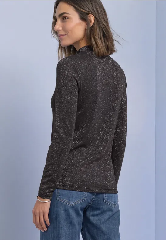 Frau steht seitlich, trägt schwarzen Glitzerpullover und blaue Jeans vor hellem Hintergrund.