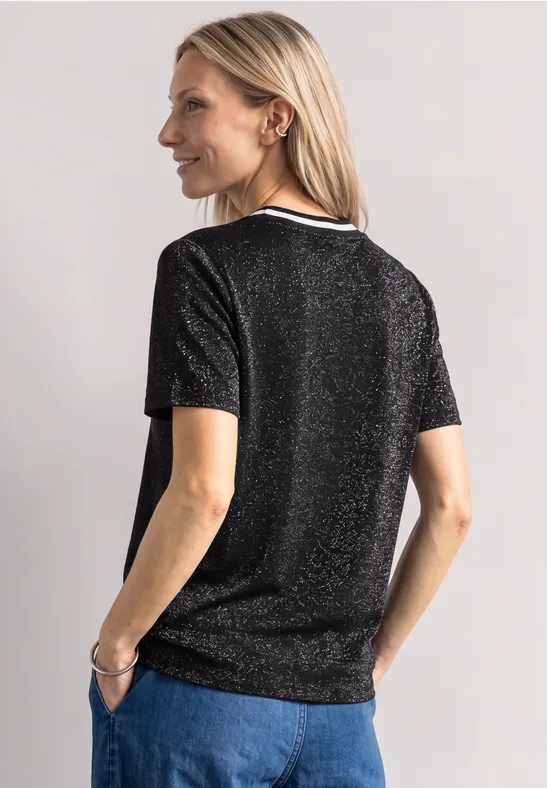 Frau von hinten in schimmerndem schwarzen T-Shirt und Jeans, schaut lächelnd über die Schulter.