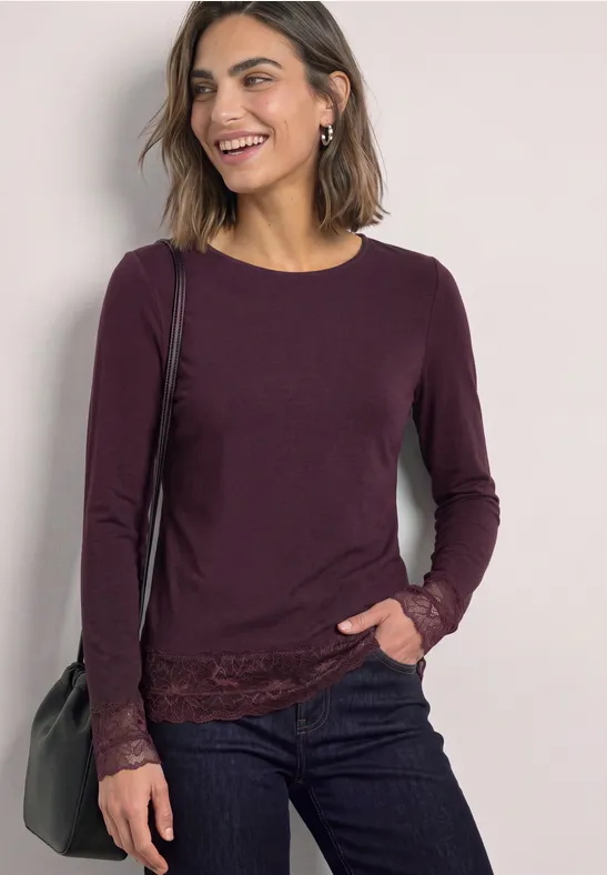 Frau in dunkellila Langarmshirt mit Spitzenbesatz an Saum und Ärmeln, dunkle Jeans, Schultertasche.