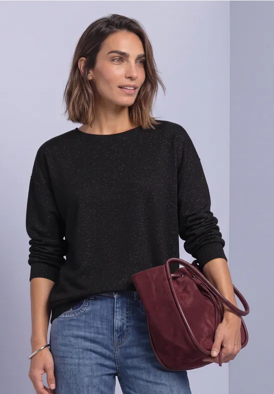 Une femme porte un pull noir, un jean et un sac bordeaux devant un fond clair.