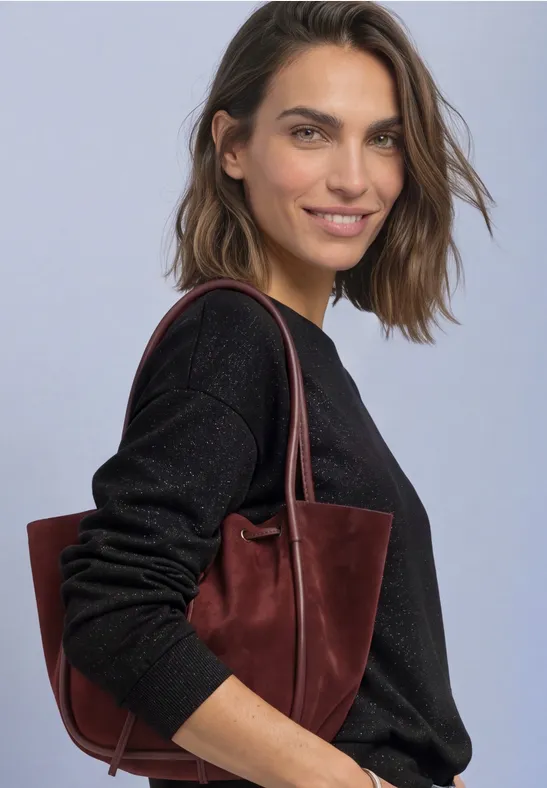 Femme souriante aux cheveux bruns porte un pull noir et un sac bordeaux sur fond clair.