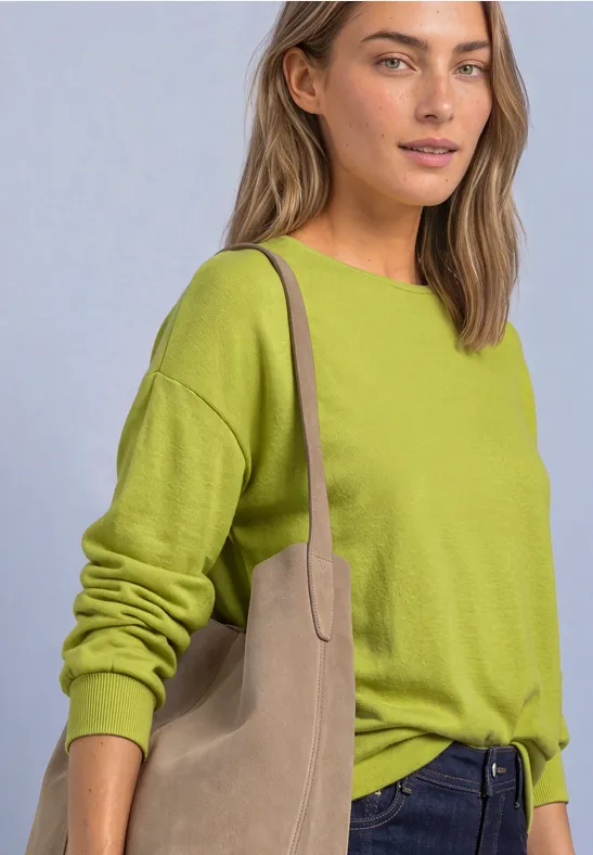 Frau in limegrünem Pullover und Jeans trägt eine beigetöne Schultertasche vor hellem Hintergrund.