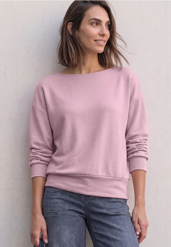 Frau steht lässig vor Wand, trägt rosa Sweatshirt und Jeans, Blick seitlich, entspannte Pose.