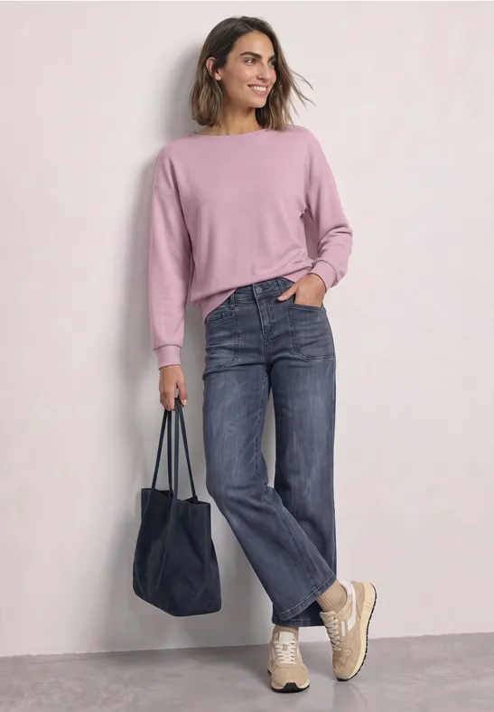 Frau in rosa Pullover, geraden Jeans und Turnschuhen trägt eine blaue Tasche und lächelt.