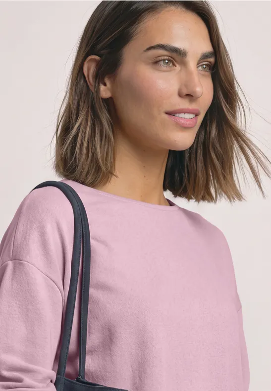 Eine junge Frau mit kurzen braunen Haaren, trägt ein rosa Shirt und eine Schultertasche.