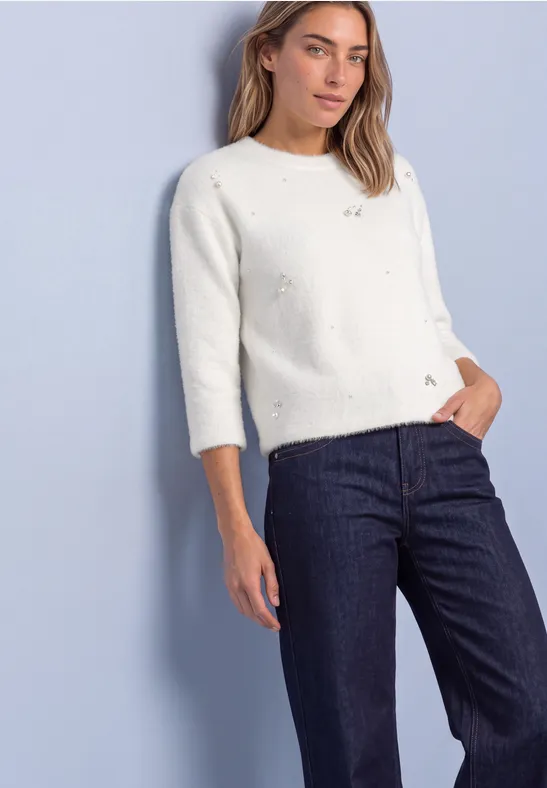 Frau trägt weißen Lochstrickpullover mit 3/4-Ärmeln und dunkle Jeans vor pastellblauem Hintergrund.