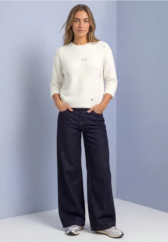 Frau in weißem Pullover und weiter, dunkler Hose, steht mit Händen in den Taschen.