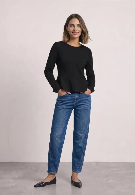 Vrouw in zwart langarmshirt, blauwe jeans en ballerina’s, glimlachend met handen in de zakken voor lichte achtergrond.