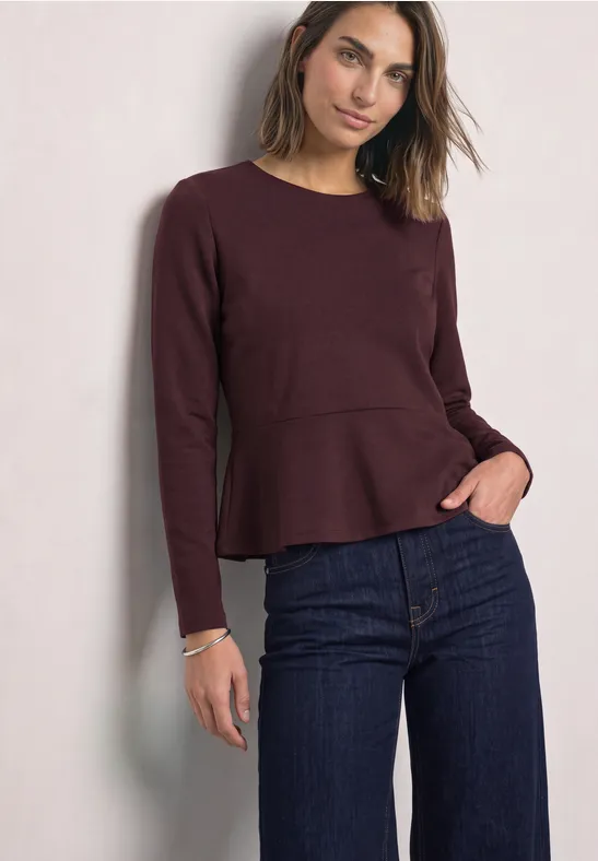 Frau in dunkelrotem Langarmshirt mit Schößchen und dunkelblauen Jeans, Hand in Tasche