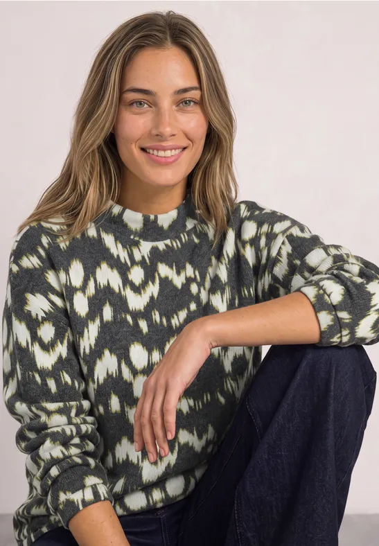 Une femme souriante assise, jambe relevée, porte un pull à motif et un pantalon sombre.