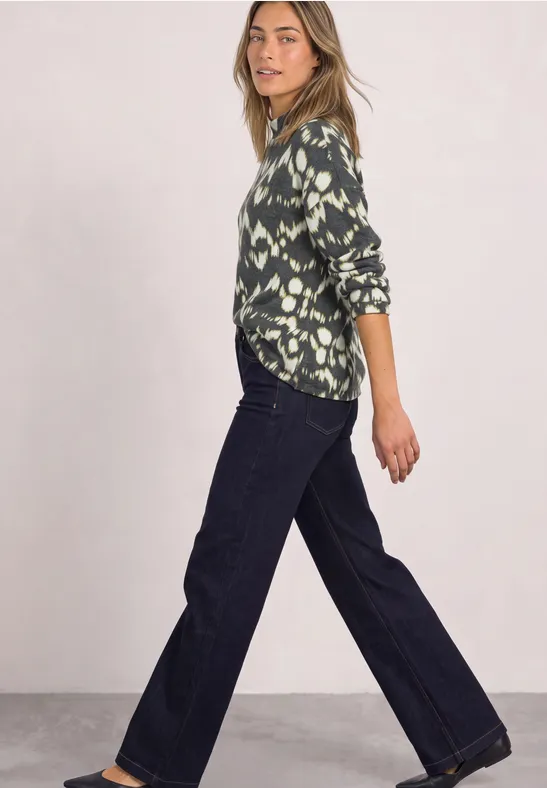 Femme en chemise verte à motifs et jean large, posée de profil devant un fond clair.