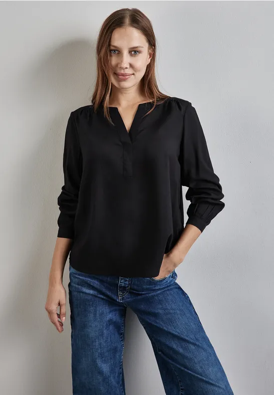 Frau mit schwarzer Bluse und blauer Jeans steht vor grauer Wand.