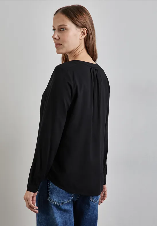 Frau mit glattem Haar trägt schwarze Bluse und Jeans, Blick über die Schulter.