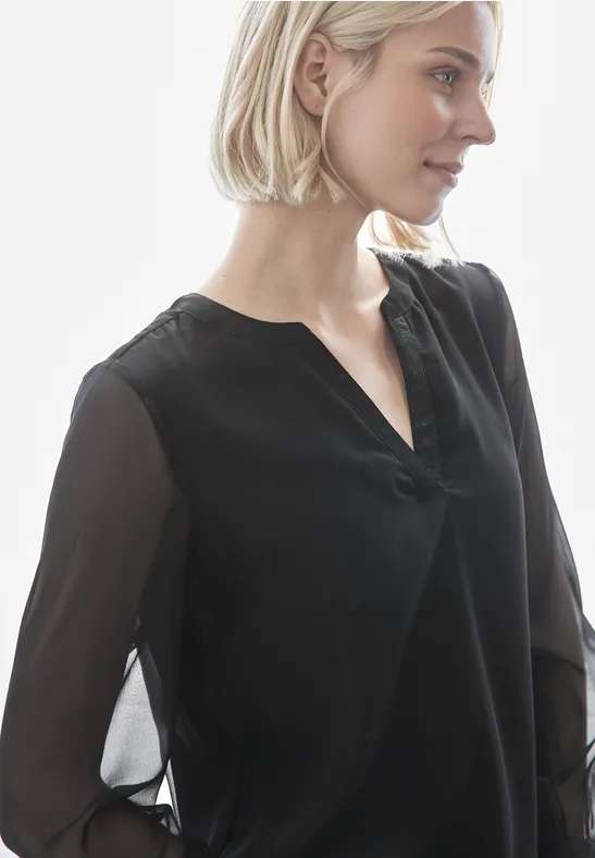 Frau mit blonden Haaren trägt eine schwarze Bluse mit transparenten Ärmeln.