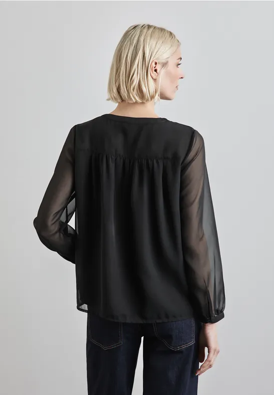 Frau mit blonden Haaren trägt schwarze, transparente Bluse und dunkle Jeans, Rückenansicht.