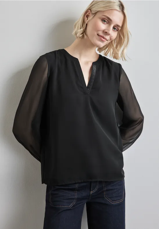 Frau mit schwarzer Bluse und dunkelblauer Jeans steht lächelnd vor einer Wand.